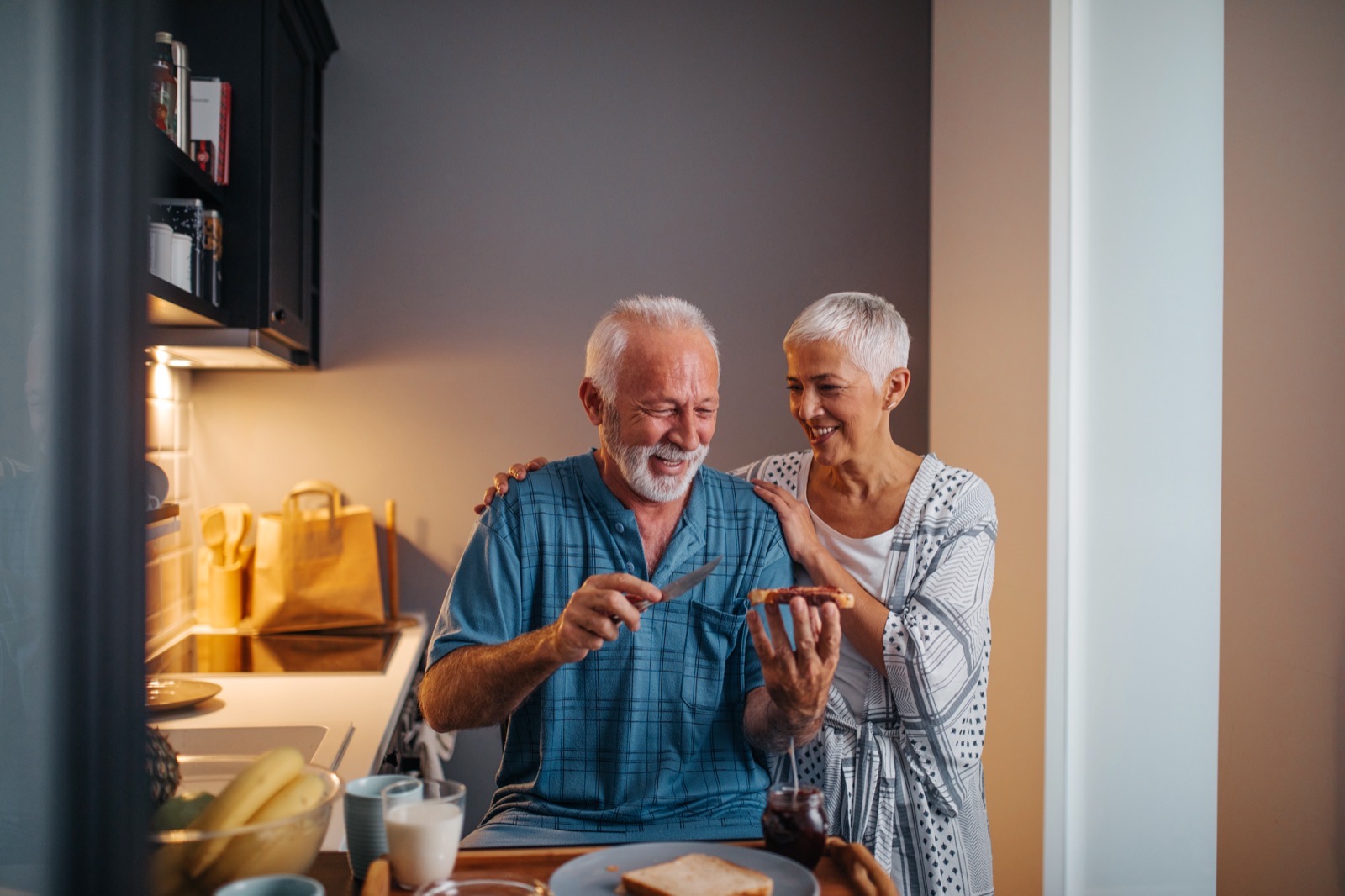 Um casal sénior ri enquanto prepara o pequeno-almoço numa cozinha cheia de luz